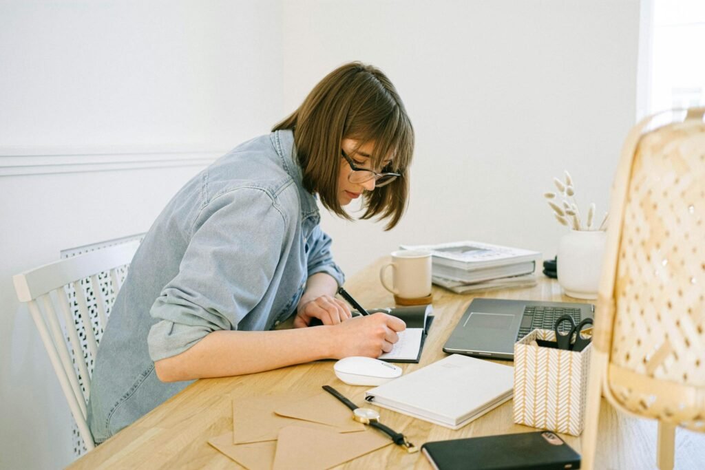 A young woman working from home, writing notes at a desk with a laptop and papers.