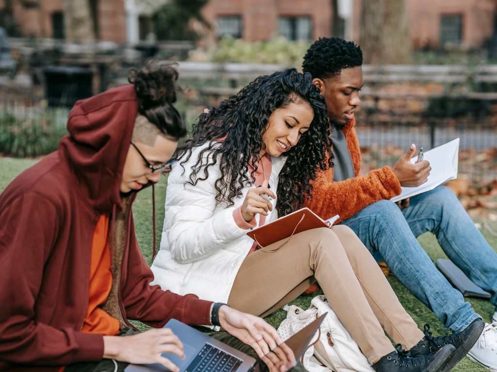 three young people reading books in a garden with one of them with a laptop