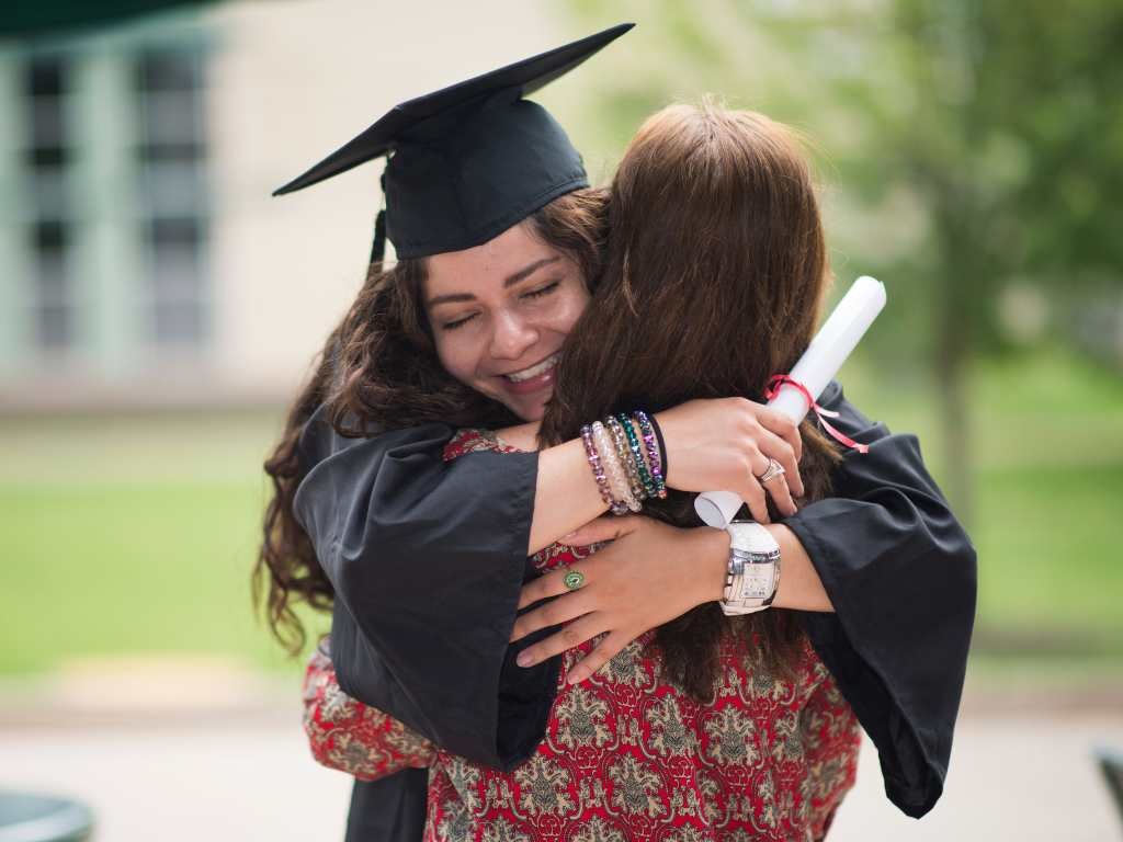 young woman who just graduated wearing a toga and a graduation cap hugging her mother with her diploma