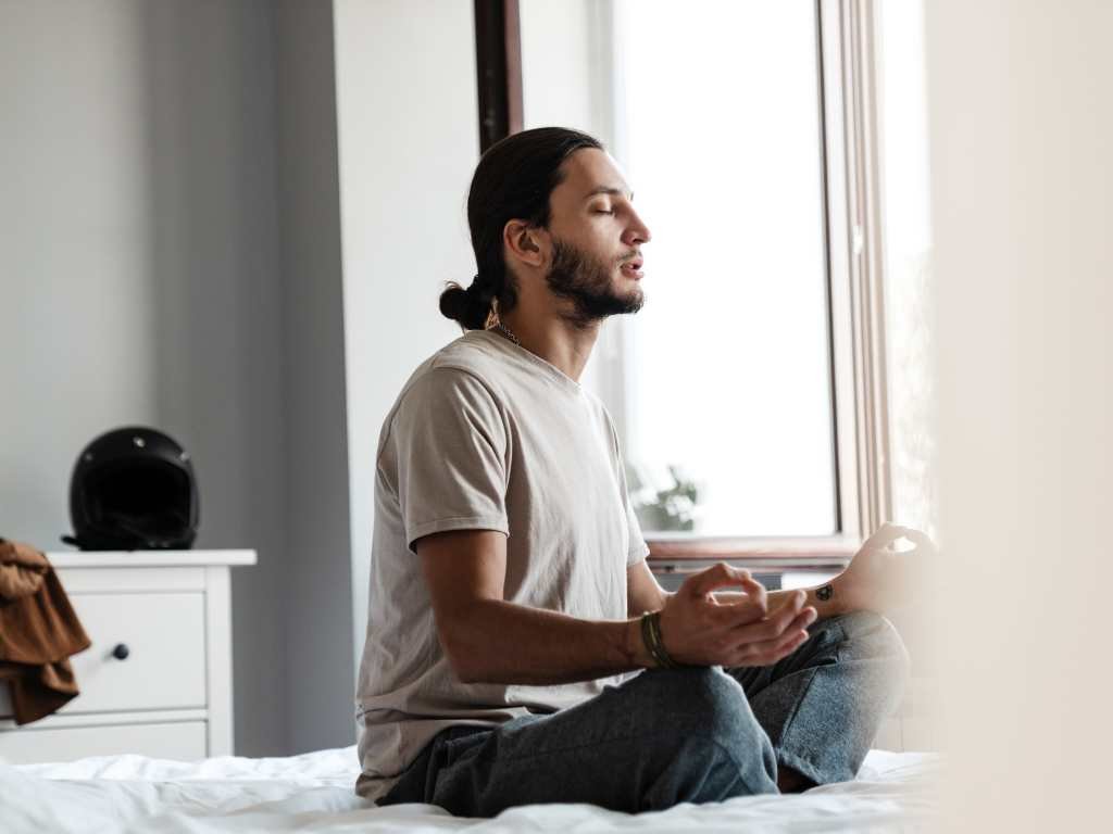 young bearded man with his hair in a ponytail meditating on top of his bed with the window open