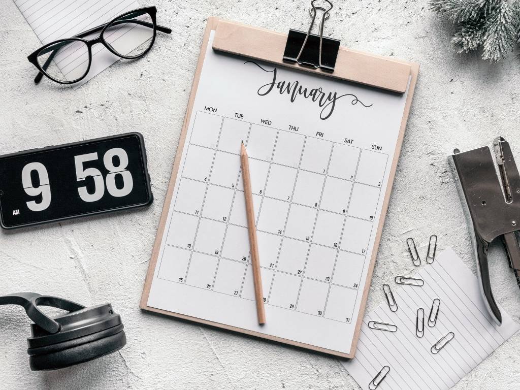A flatlay photo with a pink calendar for january with a pencil on top of it, paper clips, glasses, phone, stapler and a lid of a tumbler.