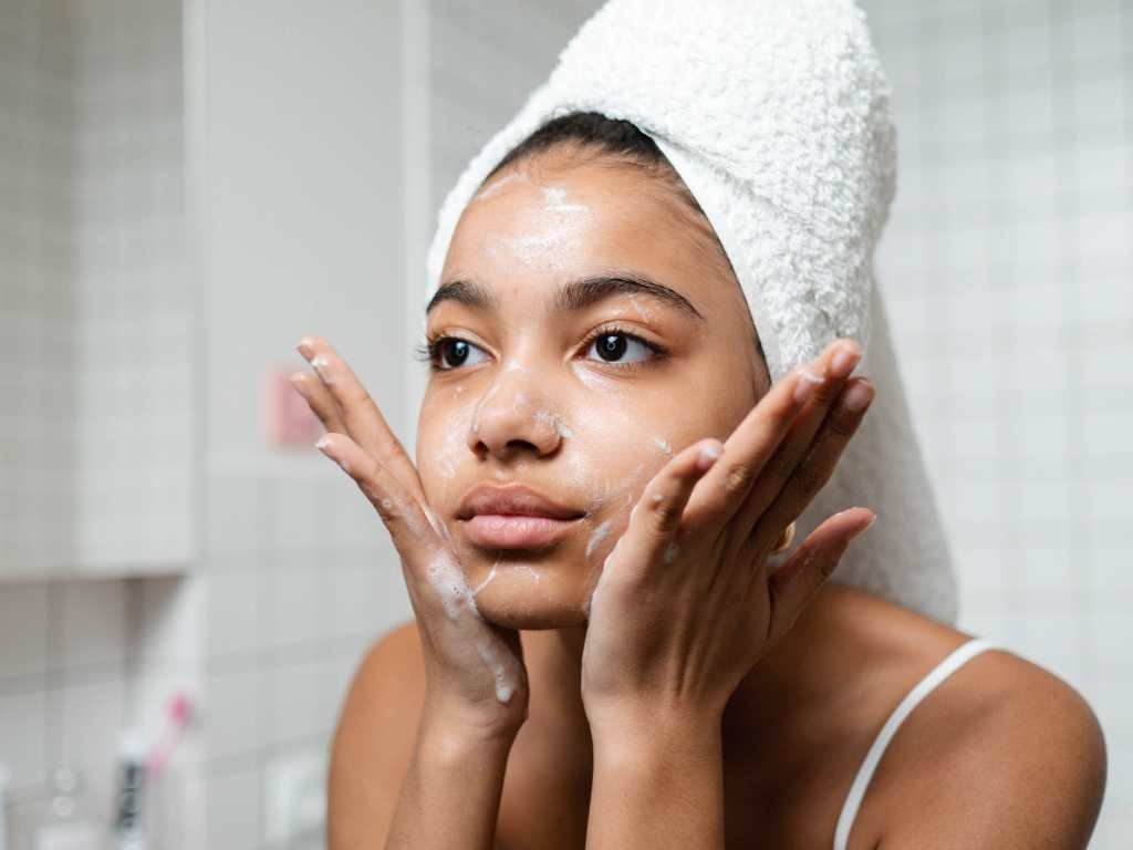 black young woman looking into a mirror with skin care cream on her face and a towel around her head