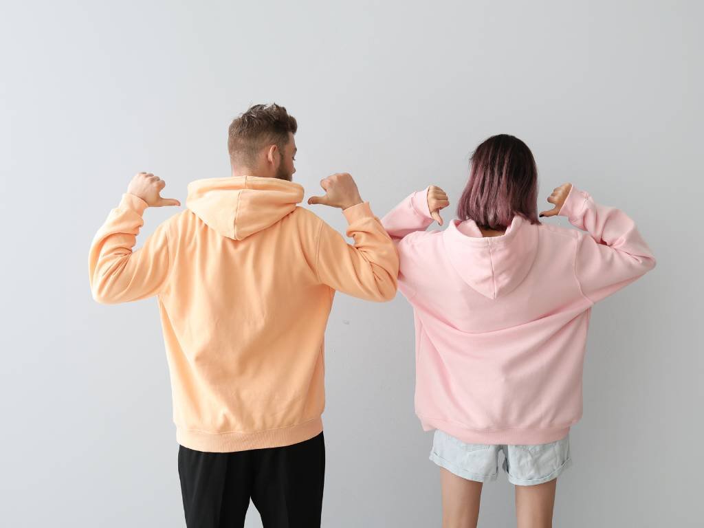 A man wearing an orange hoodie and a woman wearing a pink hoodie, both pointing to their hoodies with their thumbs