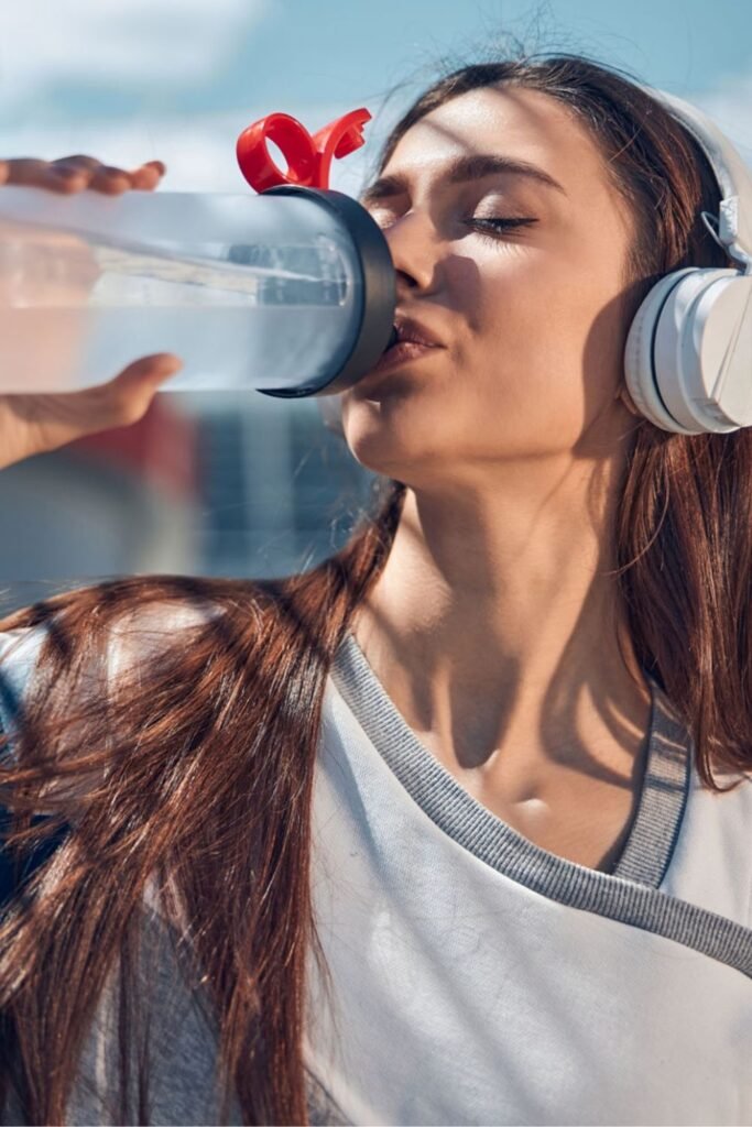 young woman drinking water from a water bottle with white headphones