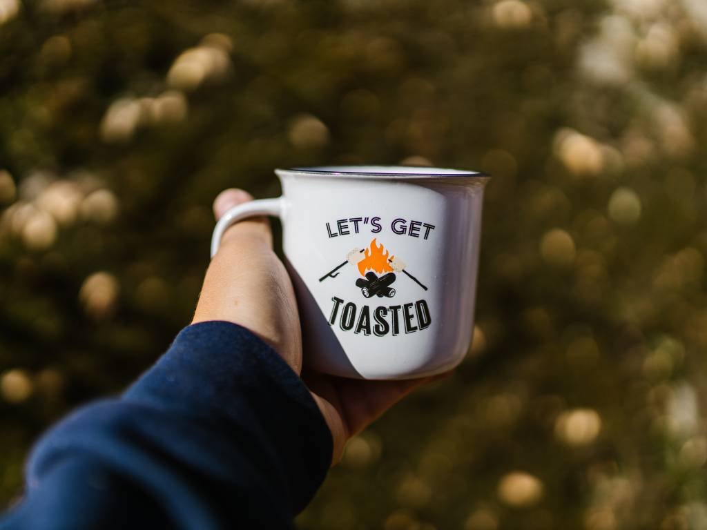 A hand wearing a blue long-sleeve shirt holding a white mug with the design 'Let's Get Roasted' on it.